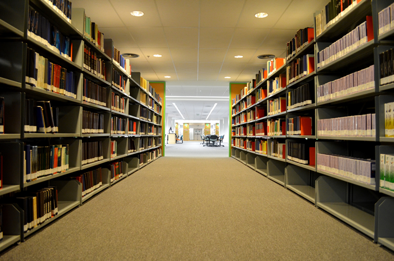 Fotografia colorida de um corredor entre duas estantes de livros da Biblioteca